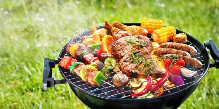 Assorted delicious grilled meat with vegetables on barbecue grill with smoke and flames in green grass